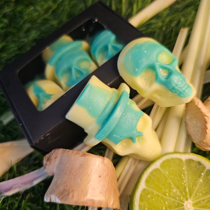 skull-shaped wax melts, with a green and yellow design, presented in a black box, surrounded by lime slices, lemongrass and ginger roots.