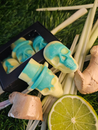 skull-shaped wax melts, with a green and yellow design, presented in a black box, surrounded by lime slices, lemongrass and ginger roots.