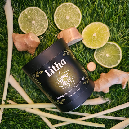 A black glass candle container labeled 'Litha', surrounded by lemongrass leaves, lime slices and ginger roots on a grassy background.