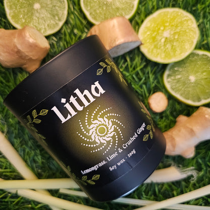 A black glass candle container labeled 'Litha', surrounded by lemongrass leaves and ginger roots on a grassy background.