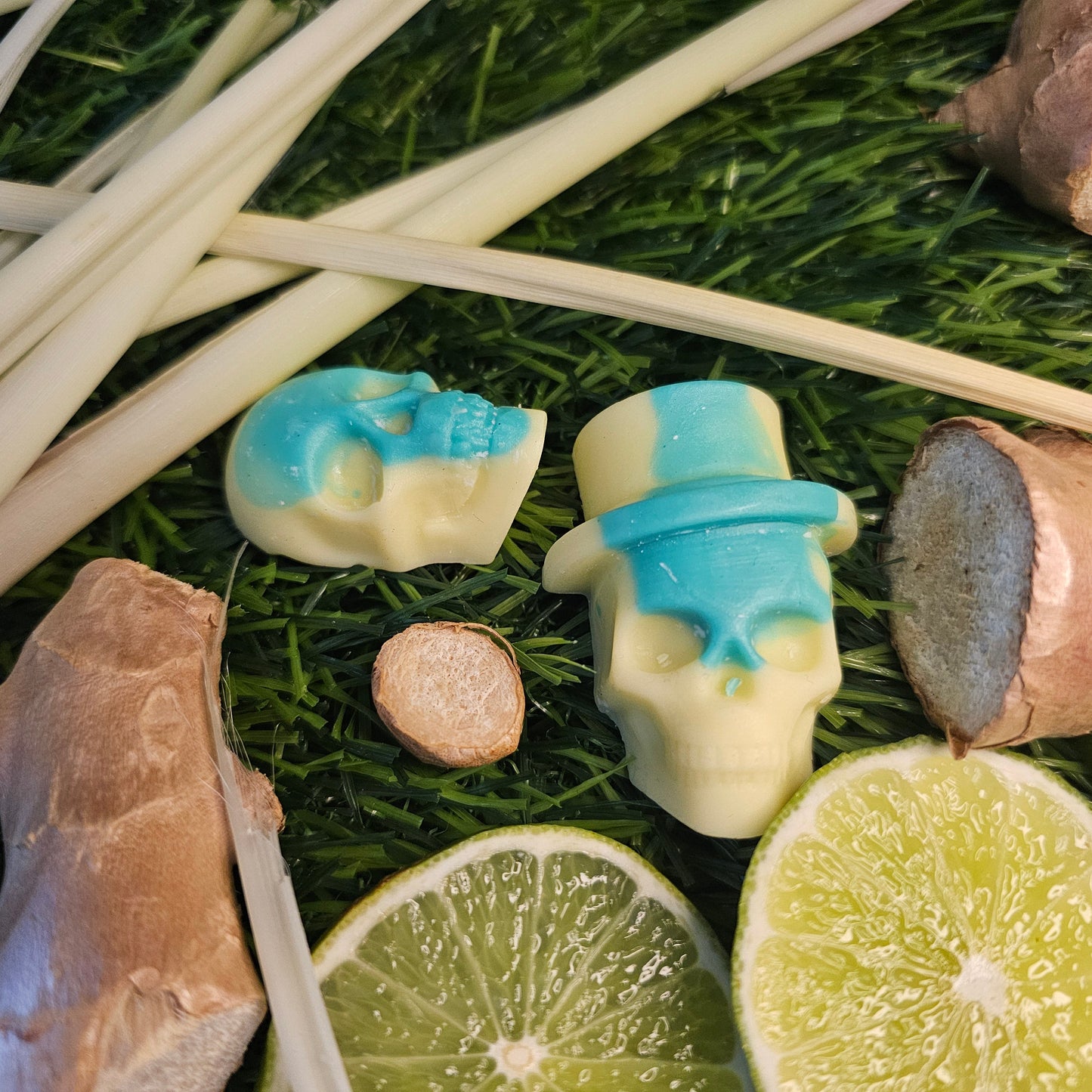 A skull-shaped wax melt, with a green and yellow design, surrounded by lime slices and ginger roots on a grassy background 