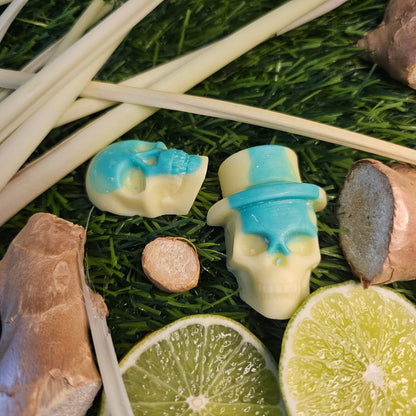 A skull-shaped wax melt, with a green and yellow design, surrounded by lime slices and ginger roots on a grassy background 