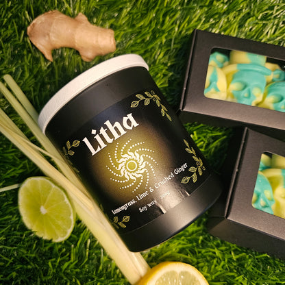 A black glass candle container labeled 'Litha', lemongrass leaves, ginger roots, lime slices and yellow and green Skull shaped wax melts lay alogside the candle on a grassy background. 