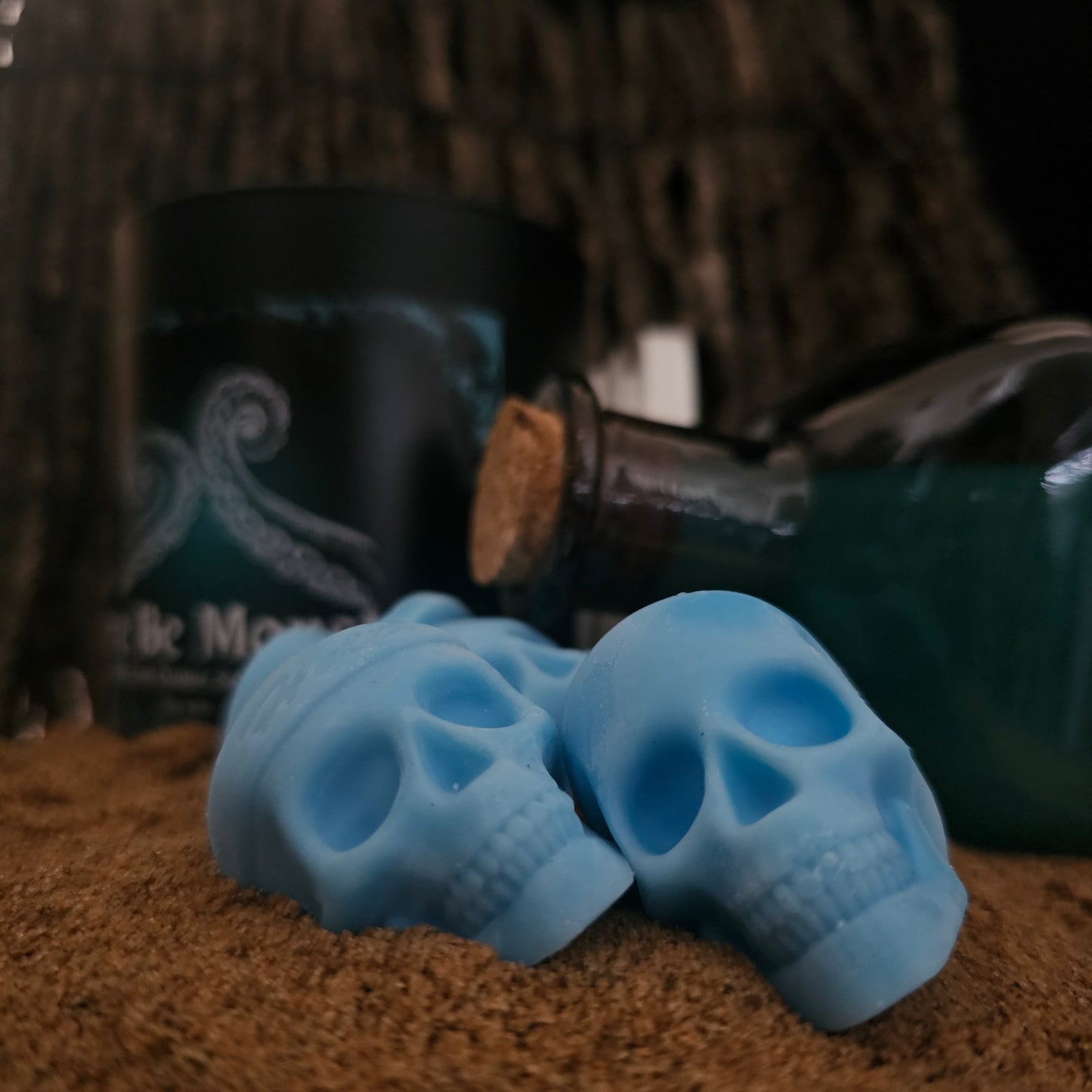 Here be monsters skull shaoed wax melts laying on a sandy surface with a here be monsters candle and blue bottle  in the background 