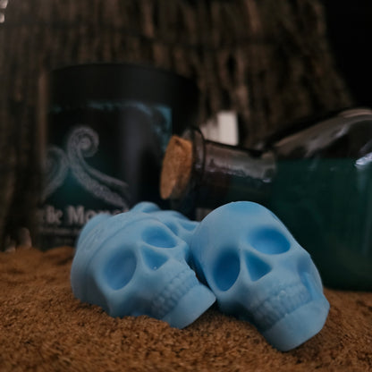 Here be monsters skull shaoed wax melts laying on a sandy surface with a here be monsters candle and blue bottle  in the background 