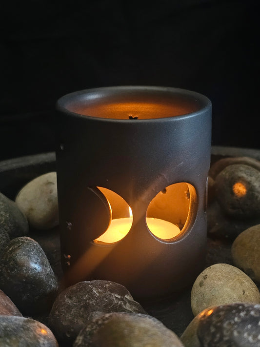 Candle in a black ceramic holder with moon phases cut out.  surrounded by stones on a dark background