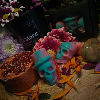 A set of green and Pink skull-shaped wax melts beside a collection of flowers and pomegranate and a candle of the same fragrance with the product name 'Ostara'