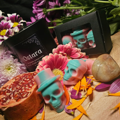 A set of green and Pink skull-shaped wax melts beside a collection of flowers and pomegranate and a candle of the same fragrance with the product name 'Ostara'