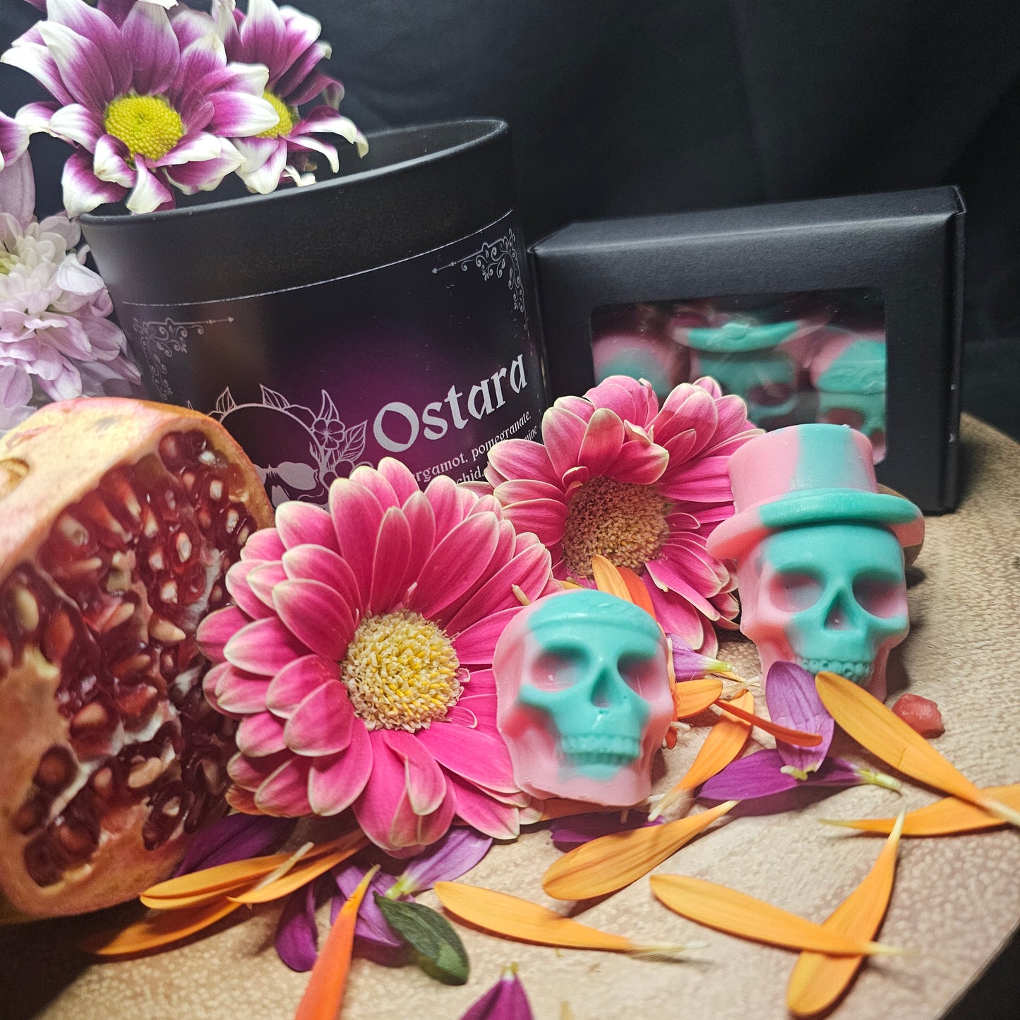 A set of green and Pink skull-shaped wax melts beside a collection of flowers and pomegranate and a candle of the same fragrance with the product name 'Ostara'