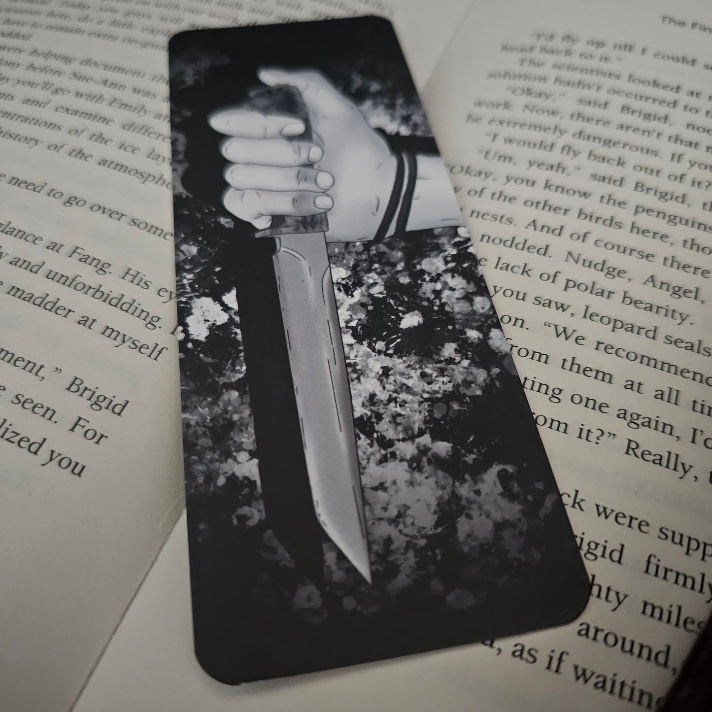 A black and white bookmark with an illustration of a hand holding a knife, placed on an open book.