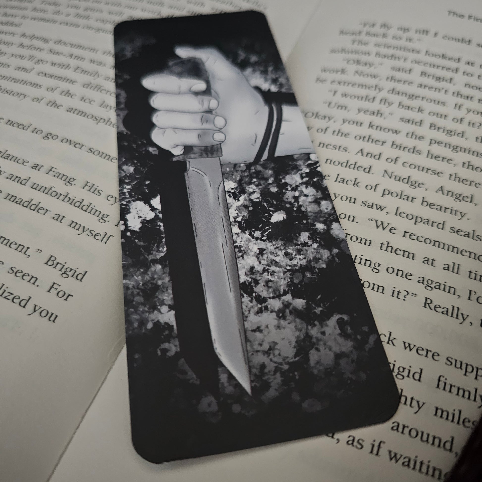 A black and white bookmark with an illustration of a hand holding a knife, placed on an open book.