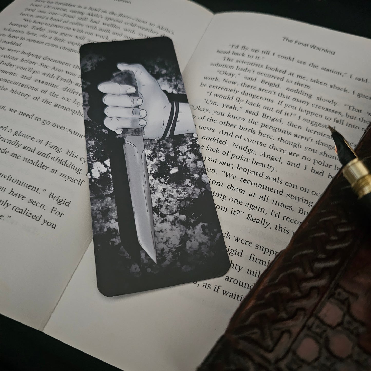 A black and white bookmark with an illustration of a hand holding a knife, placed on an open book.