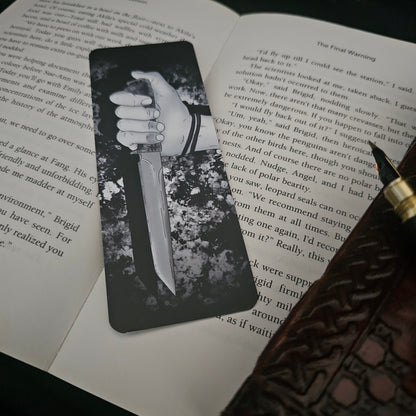 A black and white bookmark with an illustration of a hand holding a knife, placed on an open book.