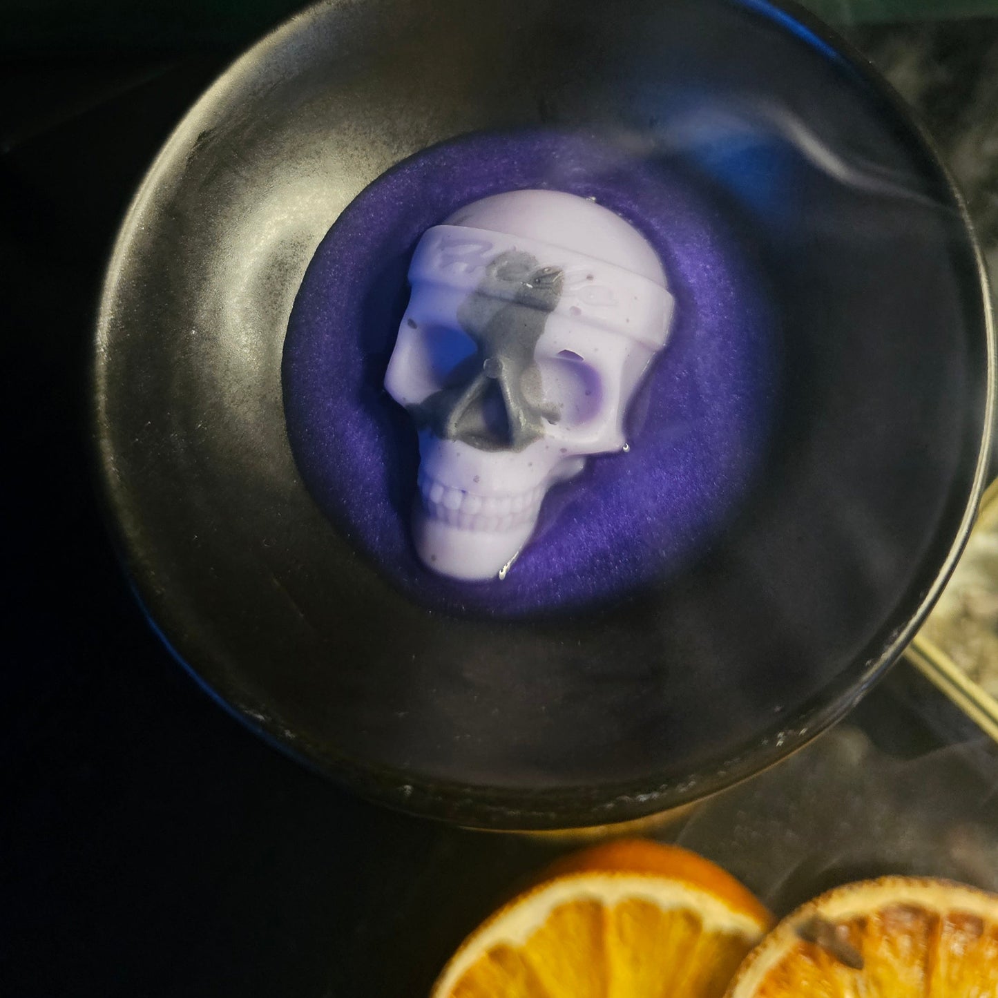 Wax melt with a skull shape in a black container, surrounded by orange slices on a dark surface.