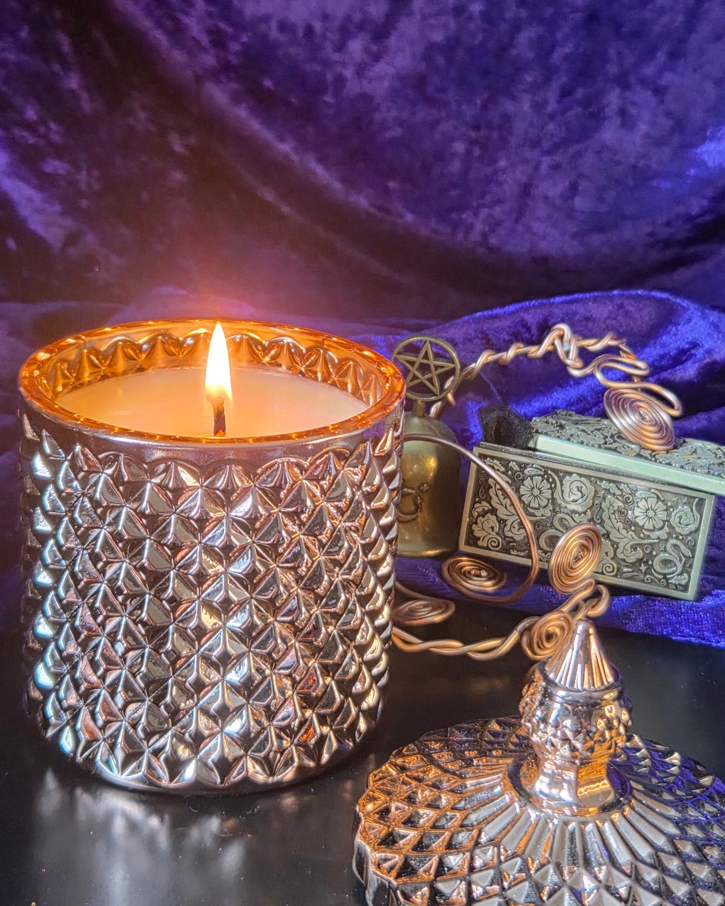 A lit candle in a textured Rose Gold glass holder sits on a dark reflective surface with its matching lid placed in front. A small decorative box and chain are visible behind it, all set against a draped purple fabric backdrop.
