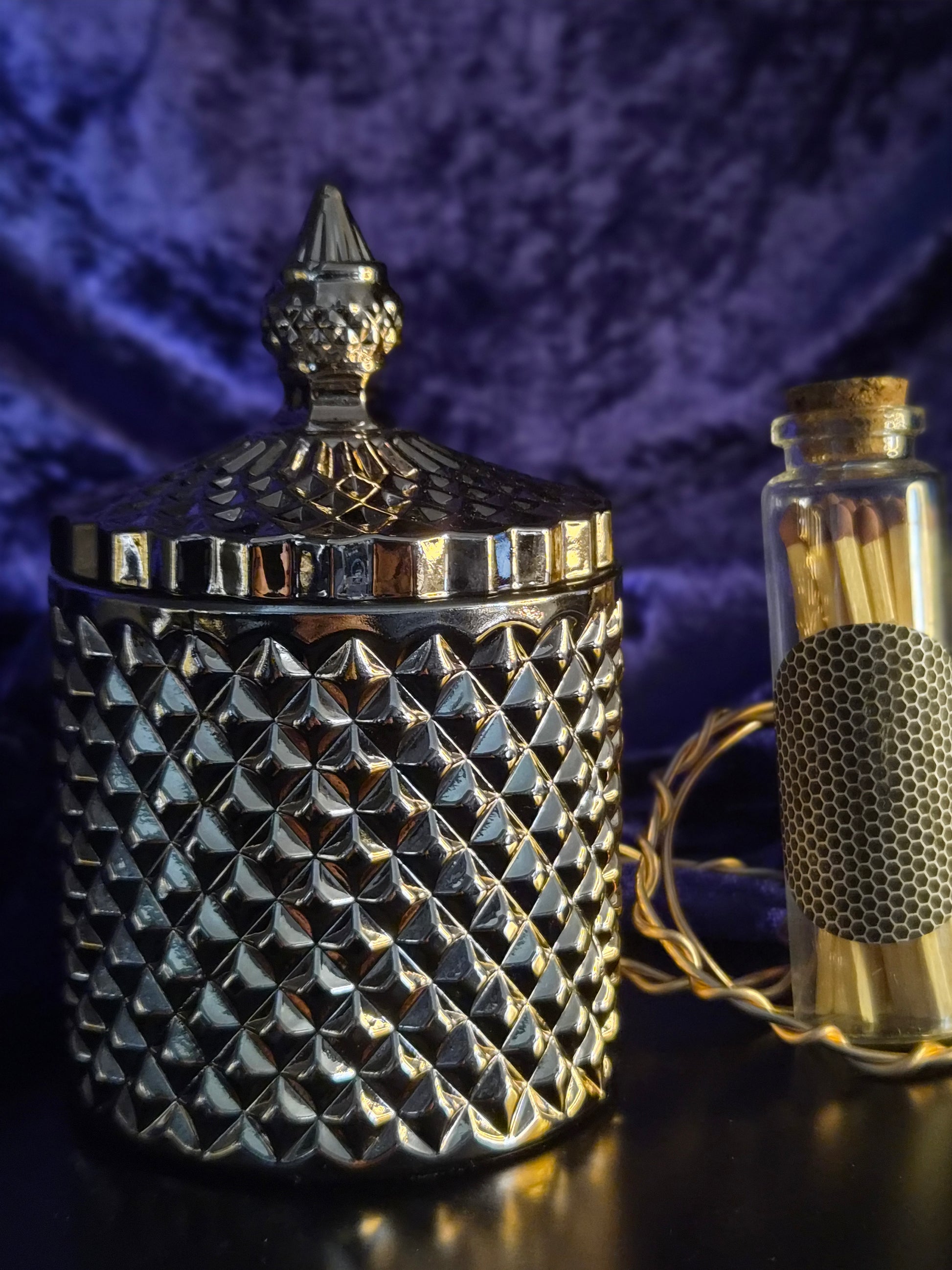 A textured lidded jar with a diamond-patterned body and pointed finial, shown next to a small glass match bottle with a cork stopper and a black mesh label, on a dark reflective surface against a purple velvet background.