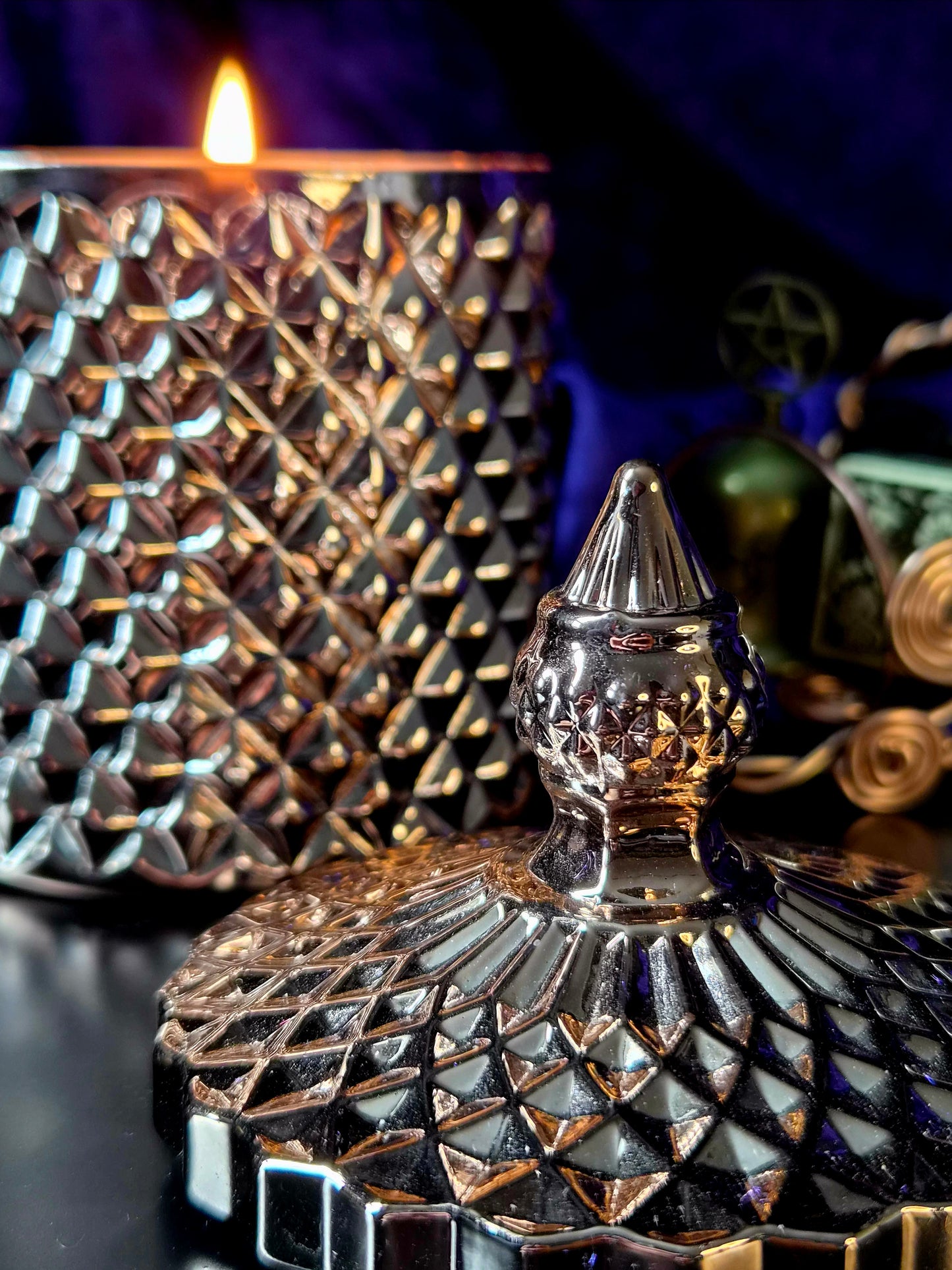 A close-up of a decorative metallic candle lid with a faceted, diamond-cut pattern and a pointed top, shown in front of a matching textured vessel with a lit candle flame. The scene is dark with warm golden light reflections and deep blue-purple background tones.