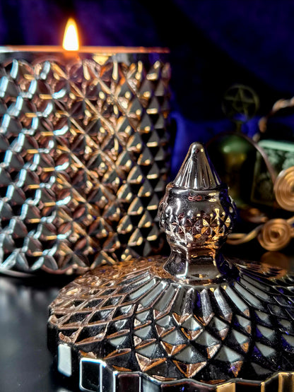 A close-up of a decorative metallic candle lid with a faceted, diamond-cut pattern and a pointed top, shown in front of a matching textured vessel with a lit candle flame. The scene is dark with warm golden light reflections and deep blue-purple background tones.
