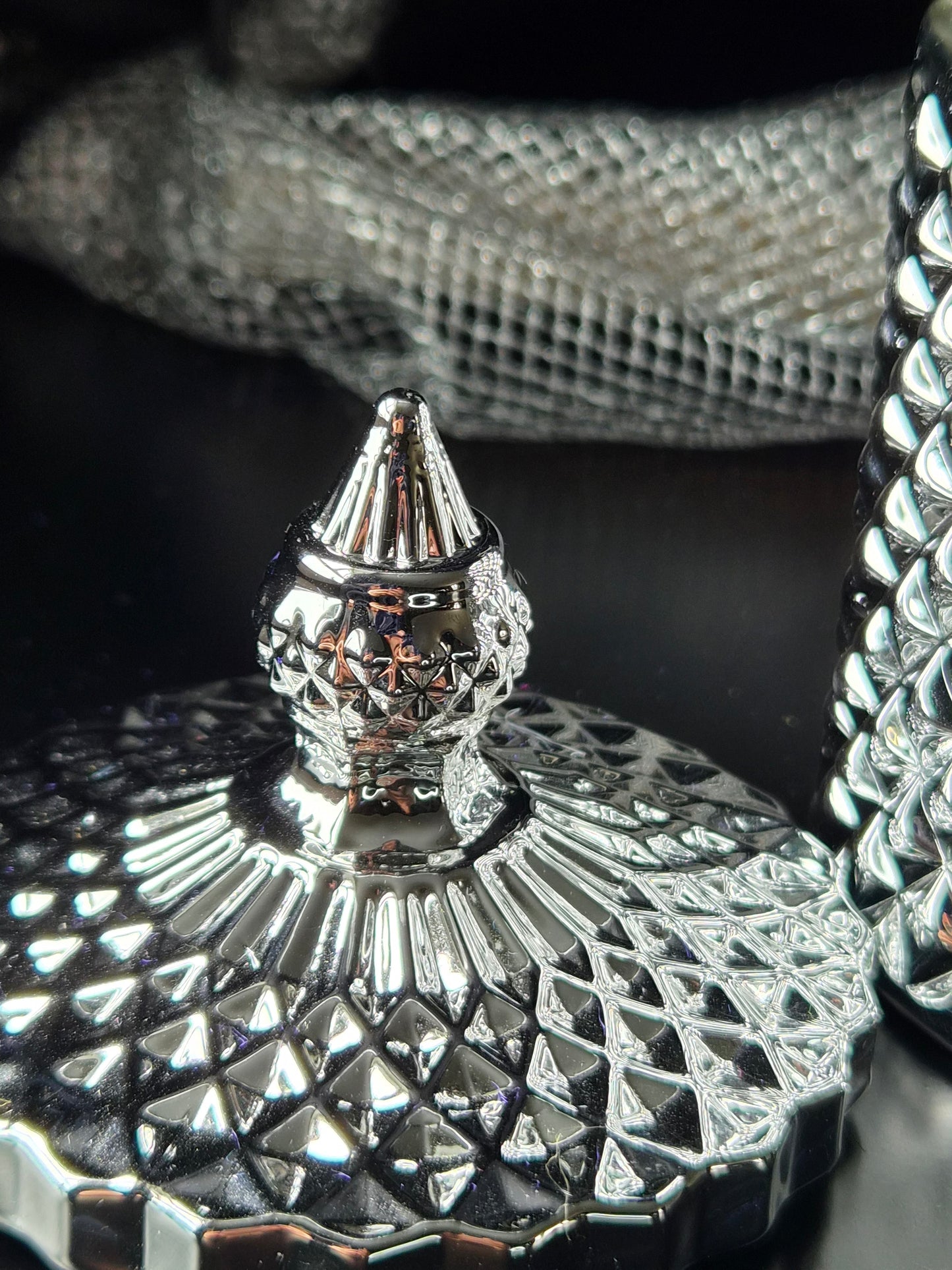 A close-up of a highly polished silver-toned  container lid with a pointed finial and a faceted diamond-cut pattern, set against a dark reflective background.
