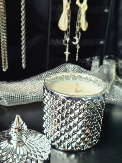 A silver textured glass candle jar with a matching lid, filled with white wax, displayed on a dark surface with jewelry and mesh fabric in the background.