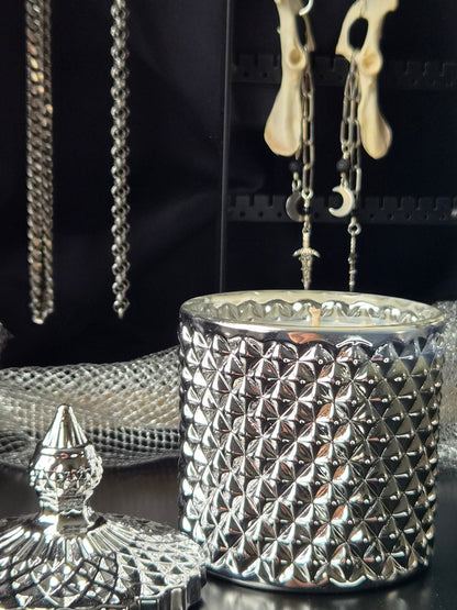 A silver textured glass jar candle with a matching lid in the foreground, set against a dark background with hanging jewelry and animal bones.