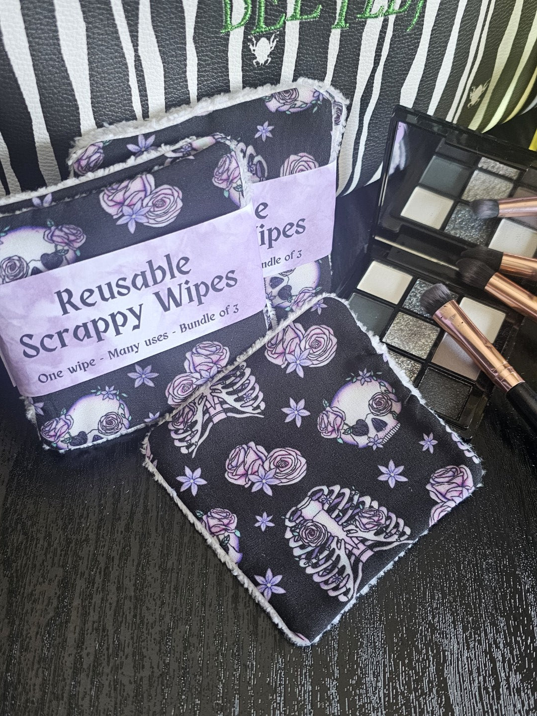 Reusable wipes with a skull, ribcage and floral design on a dark surface with makeup brushes and eyeshadow palate 
