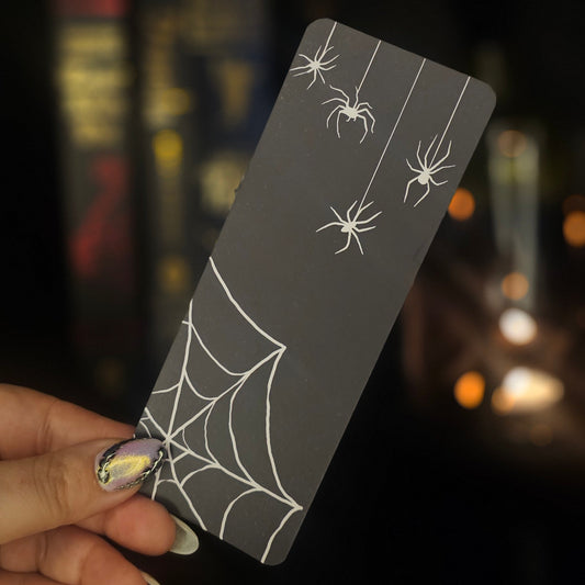 Hand holding a black bookmark with white spider web and spider designs against a blurred candlelit background.