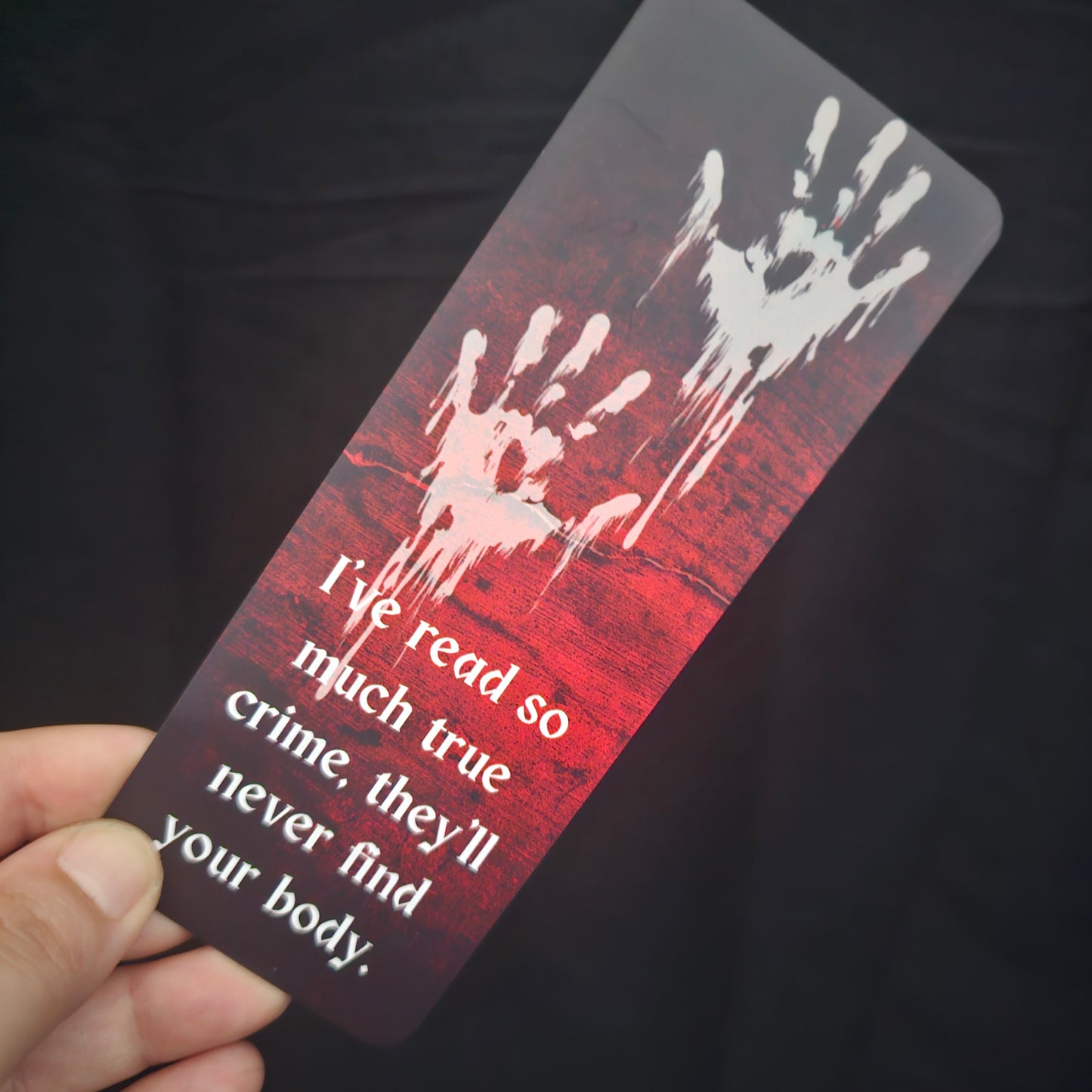 A bookmark with a blood-red background featuring handprint illustrations and the text 'I've read so much true crime, they'll never find your body.'