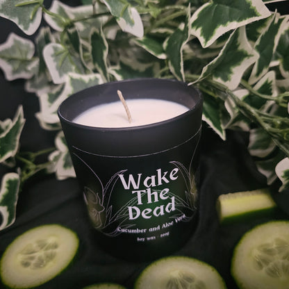 Black candle container with black and green label reading 'Wake The Dead'. Sitting on a dark surface with cucumber slices 