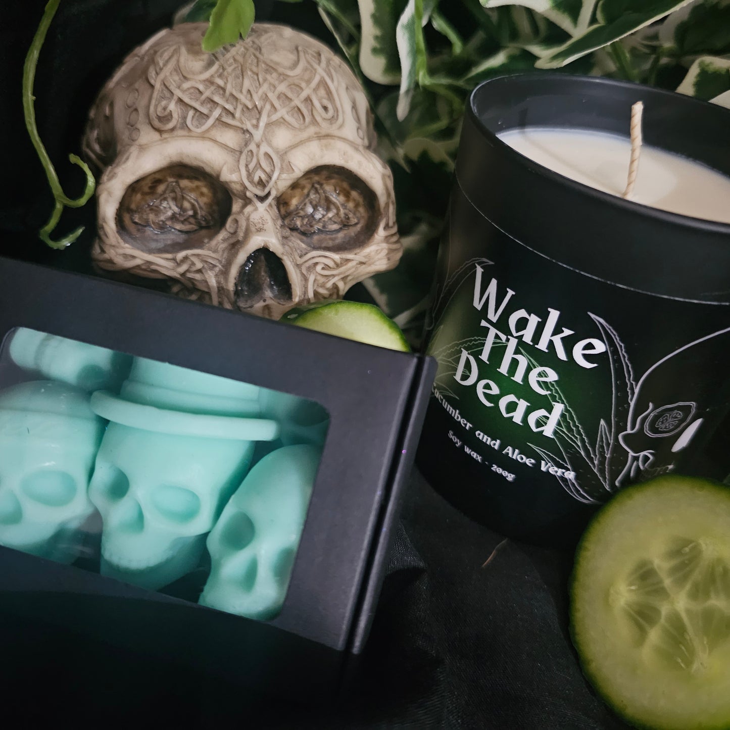 Black candle container with black and green label reading 'Wake The Dead'. Sitting on a dark surface with decorative skull, a box of skull shaped wax meltsand cucumber slices 