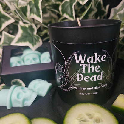 Black candle container with black and green label reading 'Wake The Dead'. Sitting on a dark surface with skull shaped wax melts and cucumber slices 