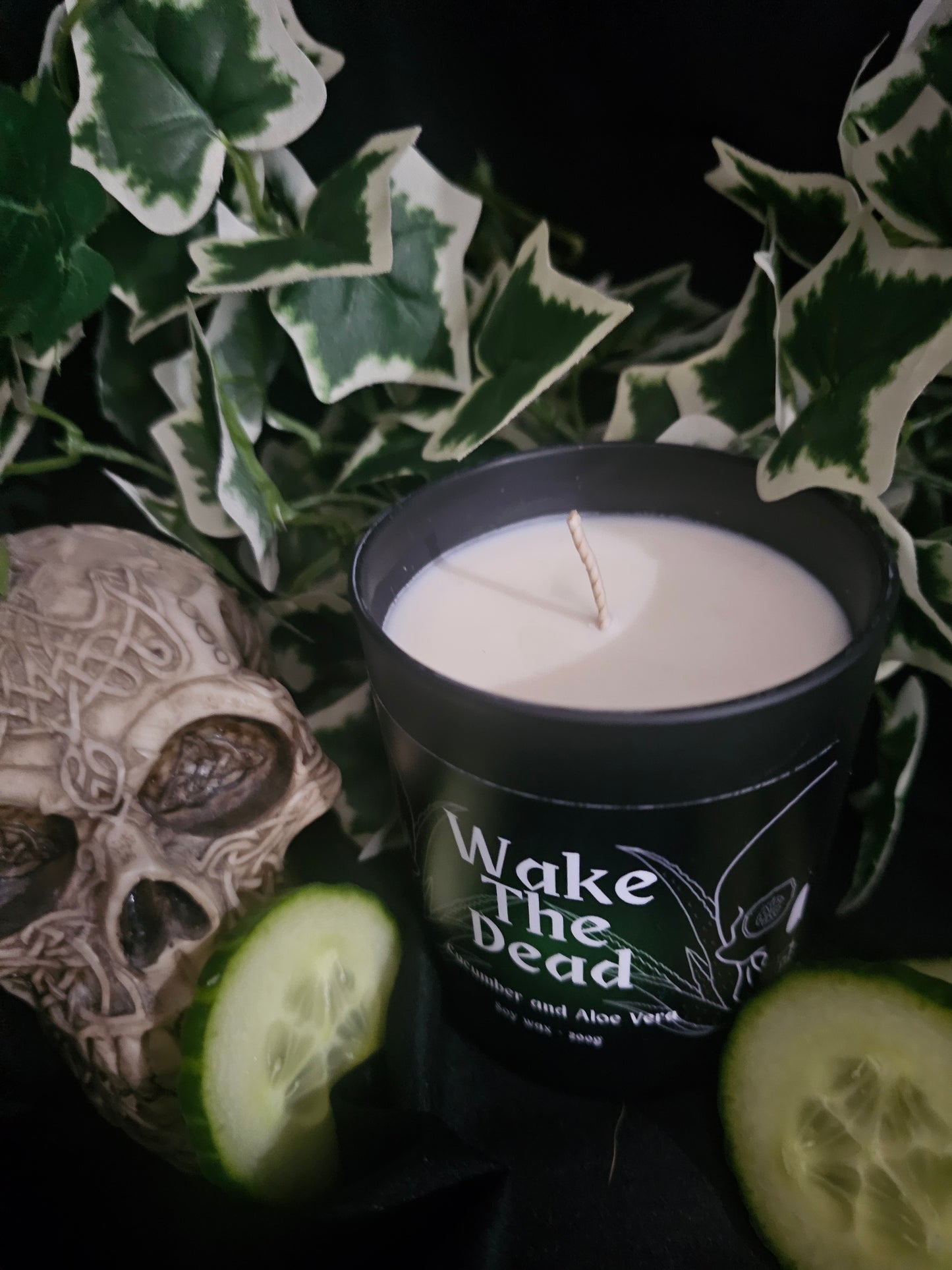 Black candle container with black and green label reading 'Wake The Dead'. Sitting on a dark surface with decorative skull and cucumber slices 