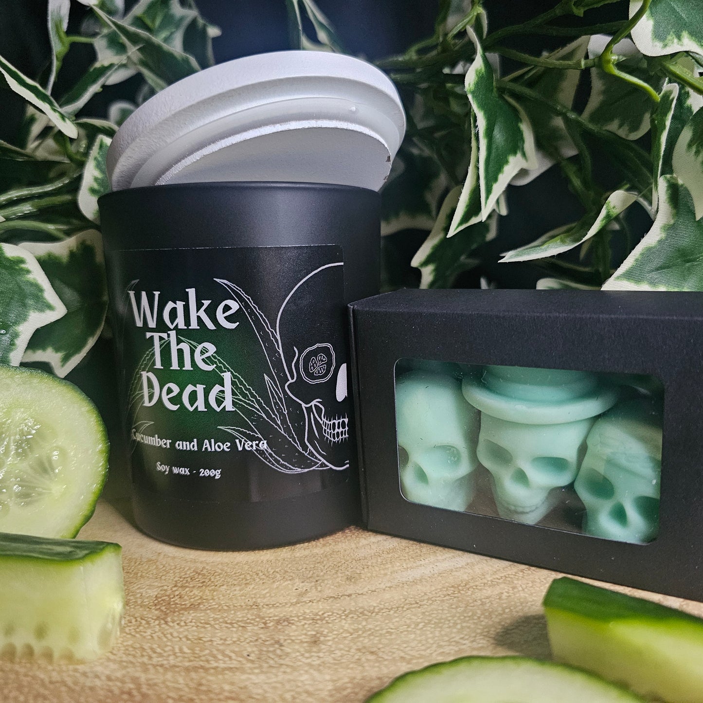 Black candle container with black and green label reading 'Wake The Dead'. Sitting on a wooden surface with skull shaped wax melts and cucumber slices 