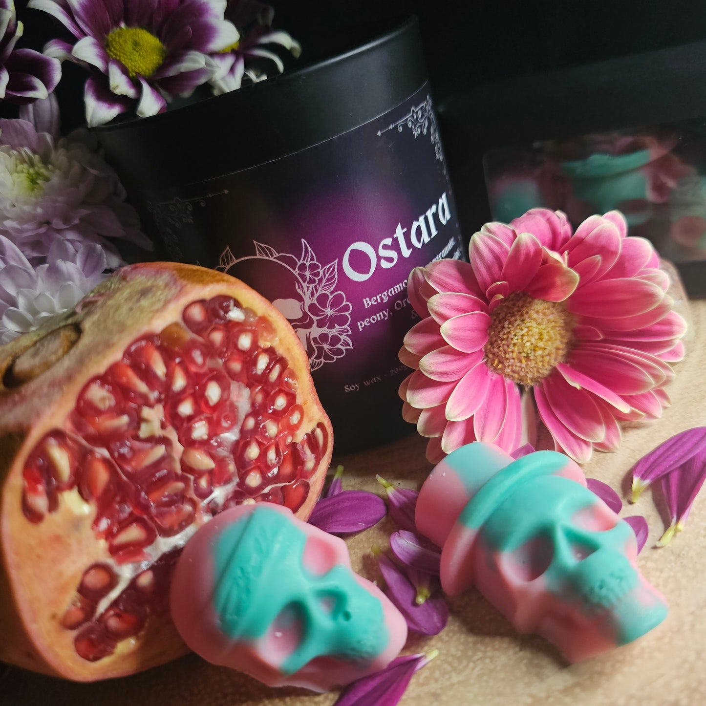 A set of green and Pink skull-shaped wax melts beside a collection of flowers and pomegranate and a candle of the same fragrance with the product name 'Ostara'