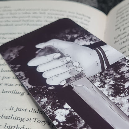 A black and white bookmark with an illustration of a hand holding a knife, placed on an open book.