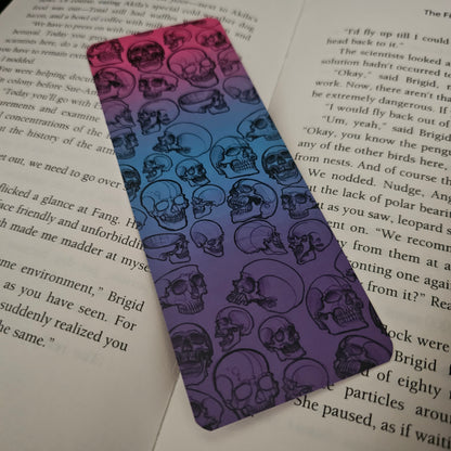 A bookmark with a gradient of pinks and blues, featuring hand-drawn skulls in various forms on it, placed on an open book.