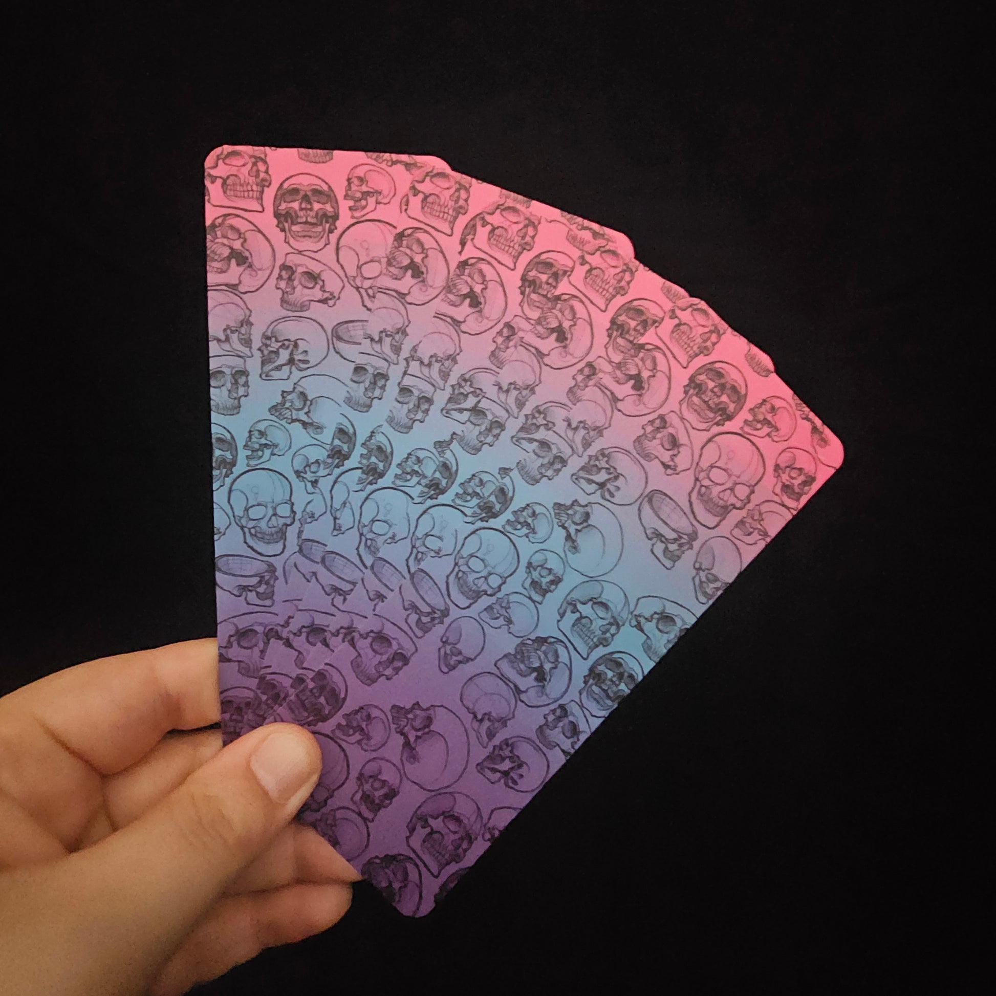 A fan of bookmarks with a gradient of pinks and blues, featuring hand-drawn skulls in various forms on a black background
