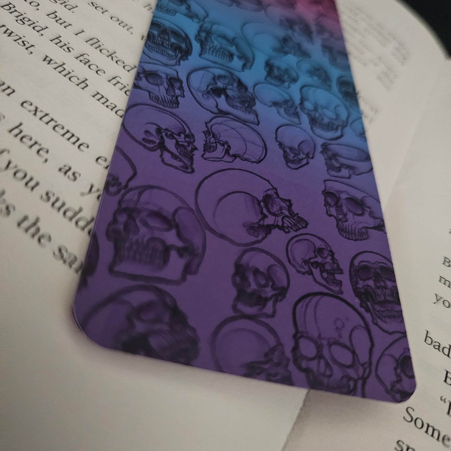 A bookmark with a gradient of pinks and blues, featuring hand-drawn skulls in various forms on it, placed on an open book, close up